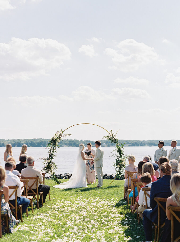 An enchanting photo of a couple exchanging vows with the serene lake and the private estate as a backdrop, surrounded by lush greenery on their special day.