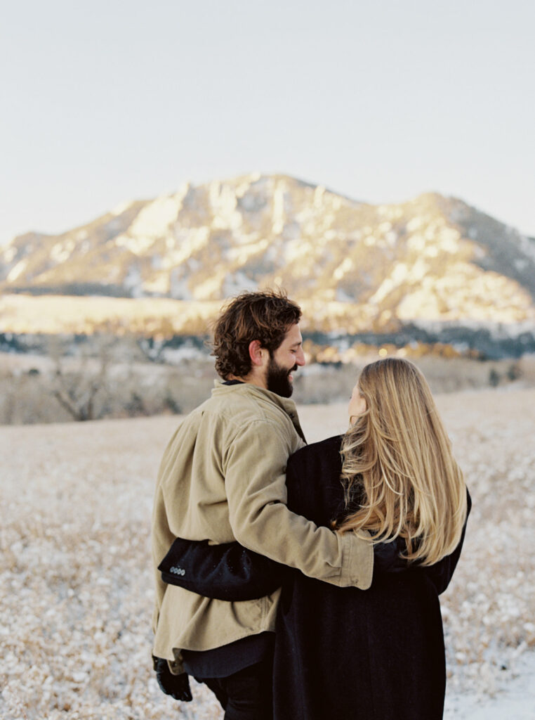 A couple stands in a snowy field, gazing into the sunrise as they embrace each other, surrounded by mountains and trees.