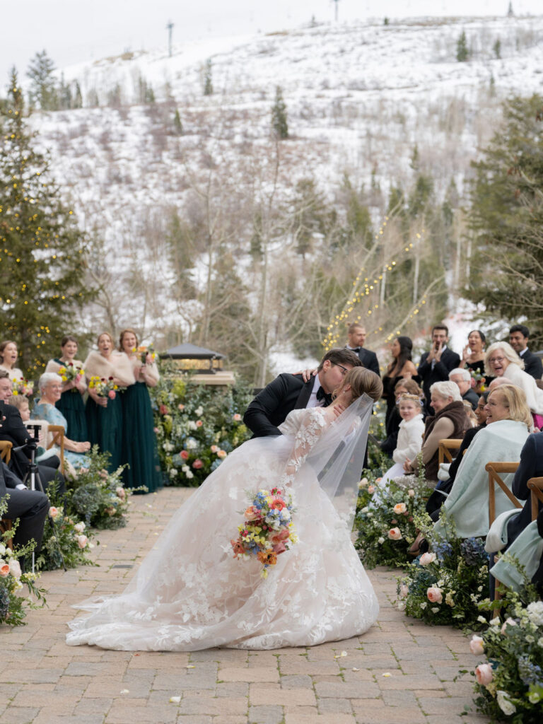 Bride and groom kiss during an outdoor winter ceremony at St. Regis Deer Valley with snow-covered mountains and guests surrounding them