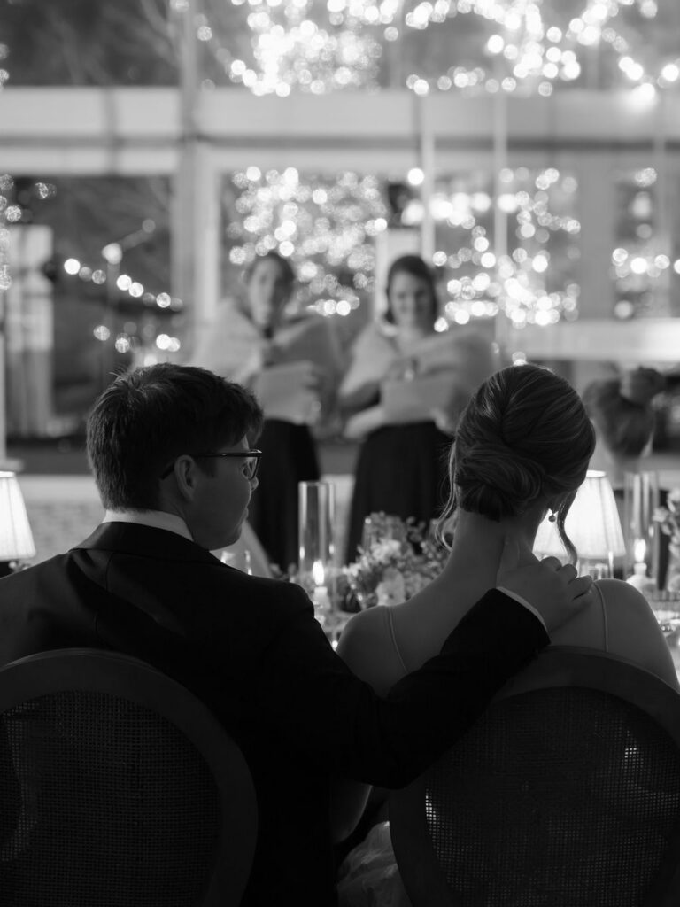 Couple seated together listening to speeches during a candlelit reception with soft string lights in the background