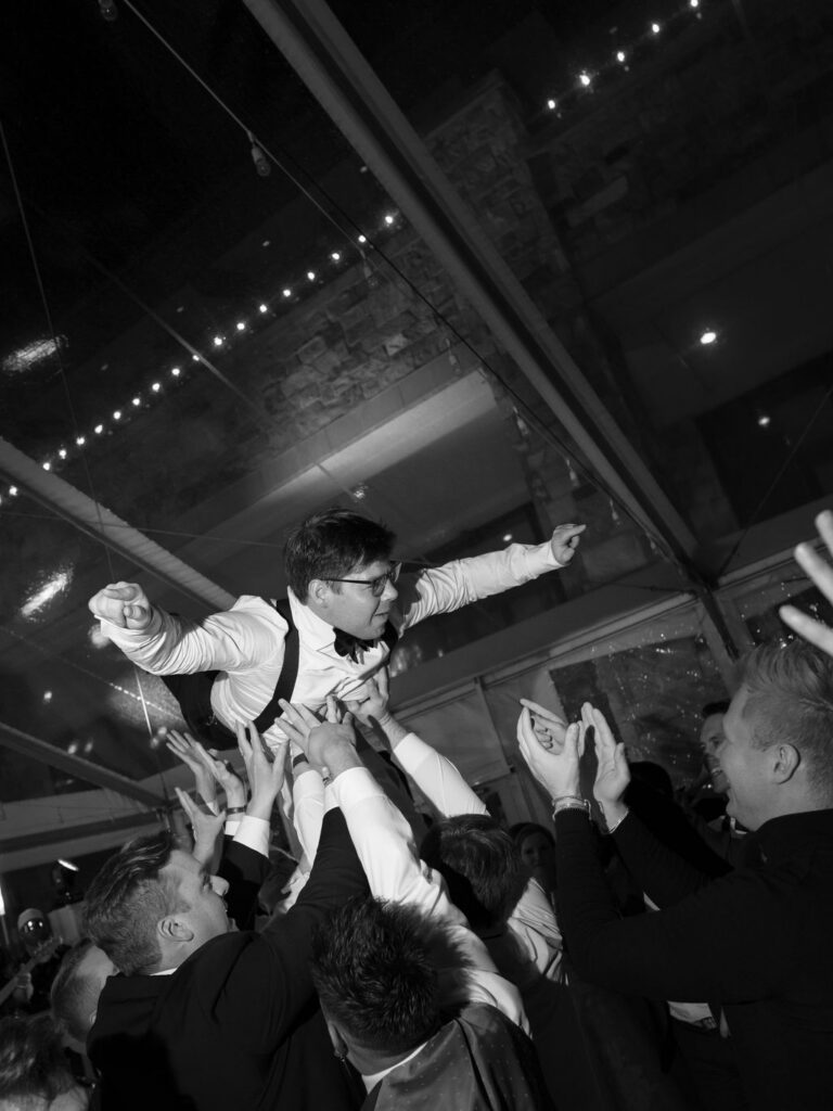 Groom being lifted in the air by guests during a lively wedding dance floor moment inside a tented reception