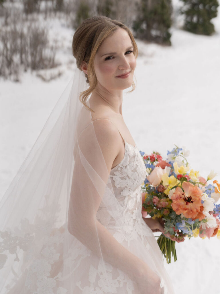 Bride standing in snowy landscape holding colorful bouquet with soft natural light in Deer Valley