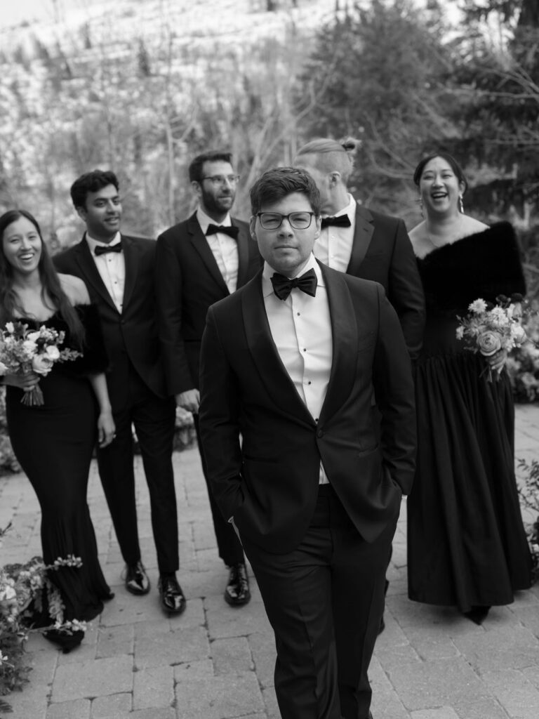 Groom walking with wedding party in formal black attire outdoors in a snowy mountain setting